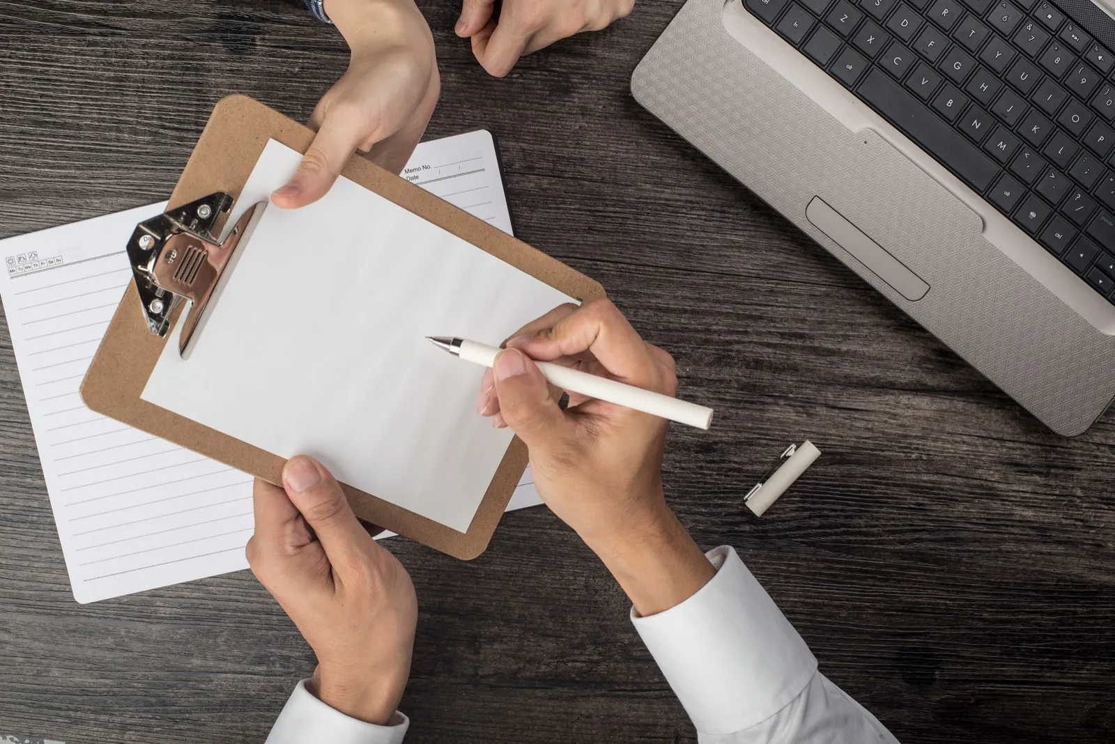 Two people at a desk, one writing on a clipboard the other holds steady.