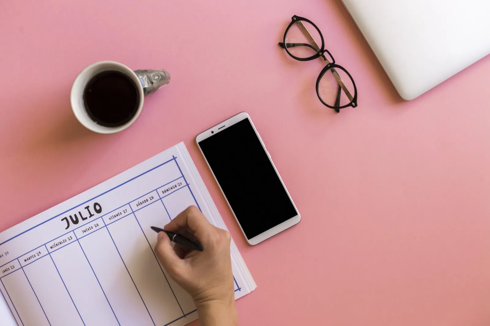 A hand fills in a paper week planner beside a phone and a cup of coffee.