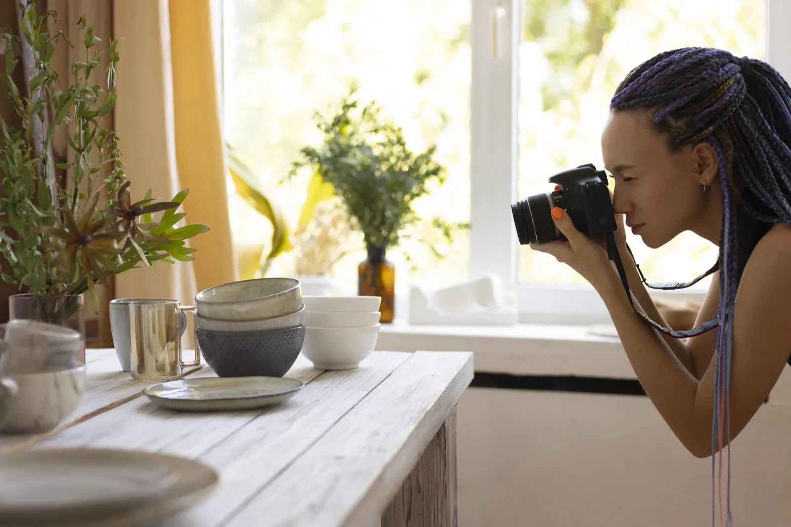 A woman photographs stacked ceramic bowls on a kitchen counter by a window.
