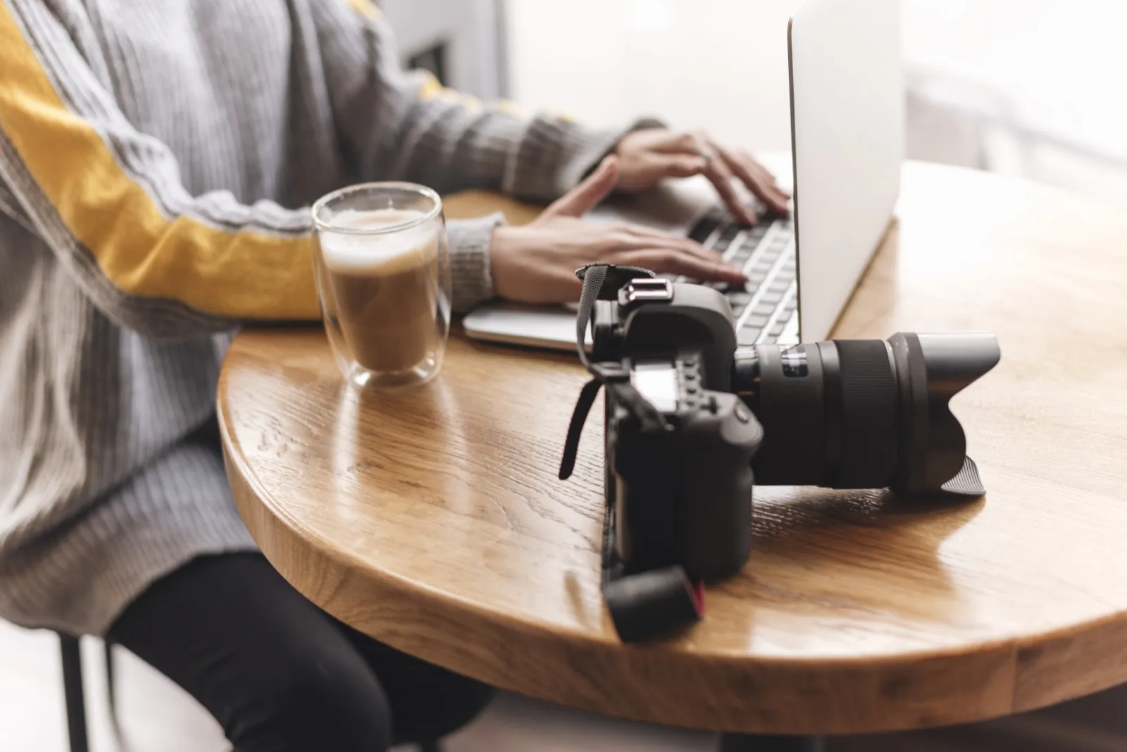 Hands typing on a laptop at a cafe table, a camera resting beside it.