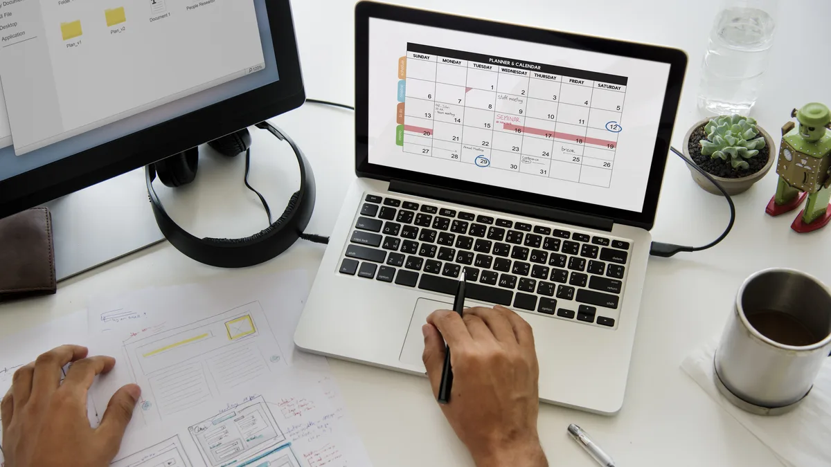 Hands at a laptop showing a monthly calendar, with printed schedules and coffee beside it.