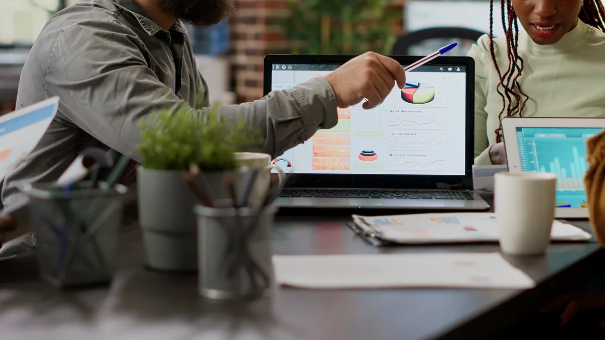 A hand pointing at charts on a laptop screen while colleagues compare printouts around it.