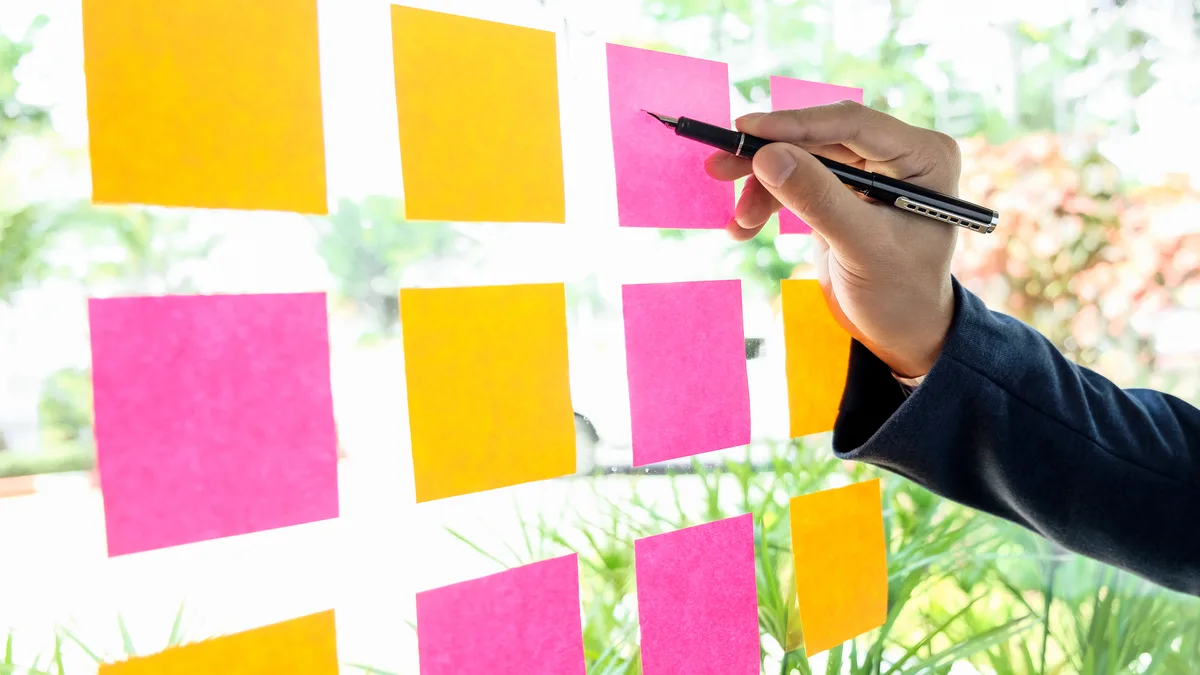 A hand adding orange and pink sticky notes to a grid on an office glass wall.