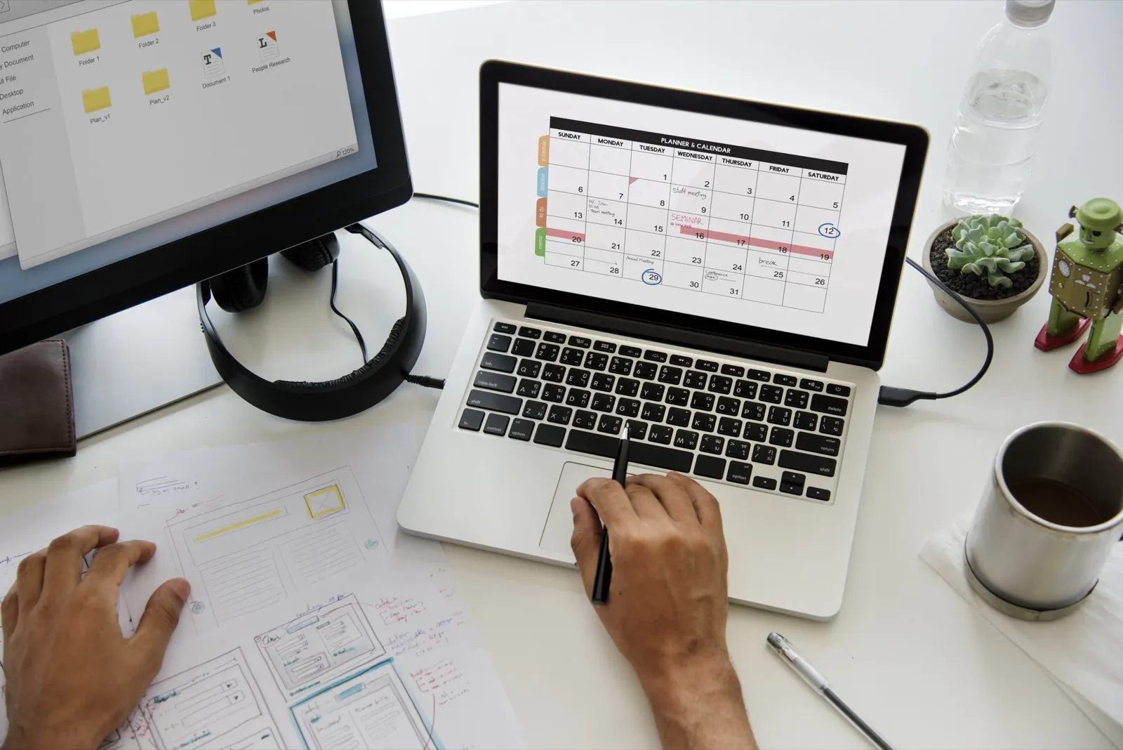 Hands at a laptop showing a month's planner calendar, printed layouts marked up beside it.