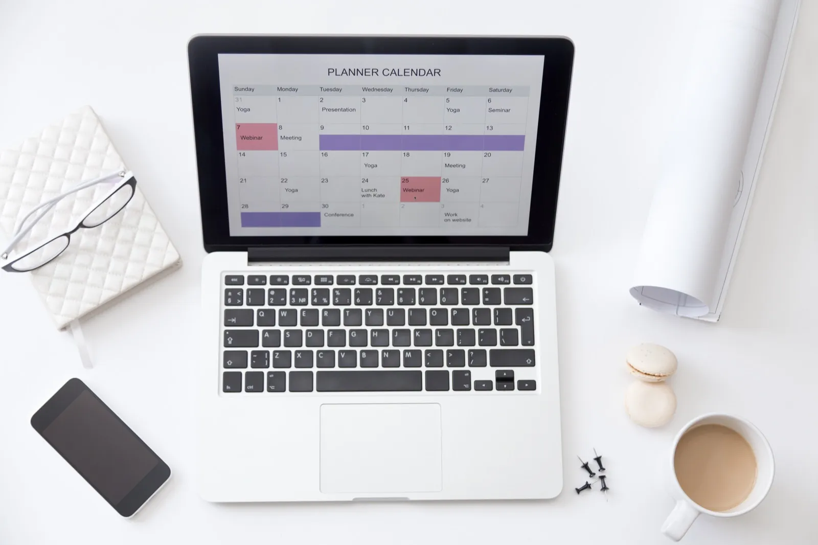 A laptop showing a month's planner calendar, seen from above beside a phone and notebook.