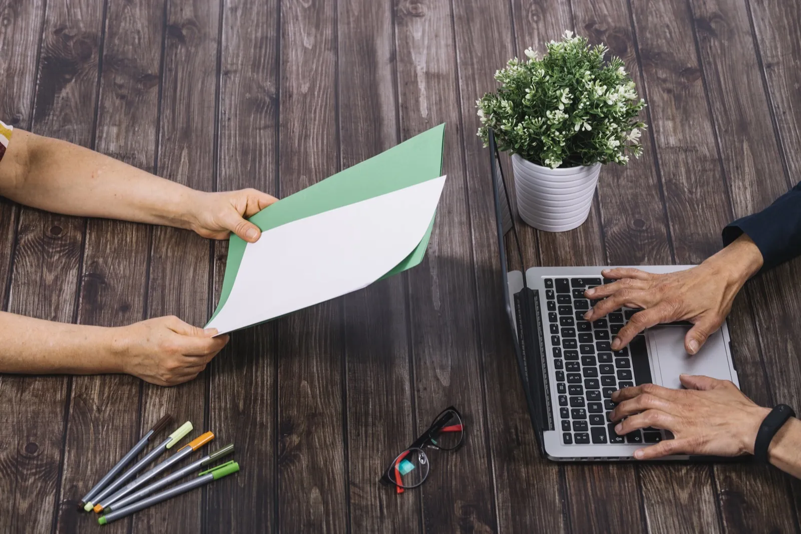 One person hands a printed sheet across a desk while another types on a laptop.