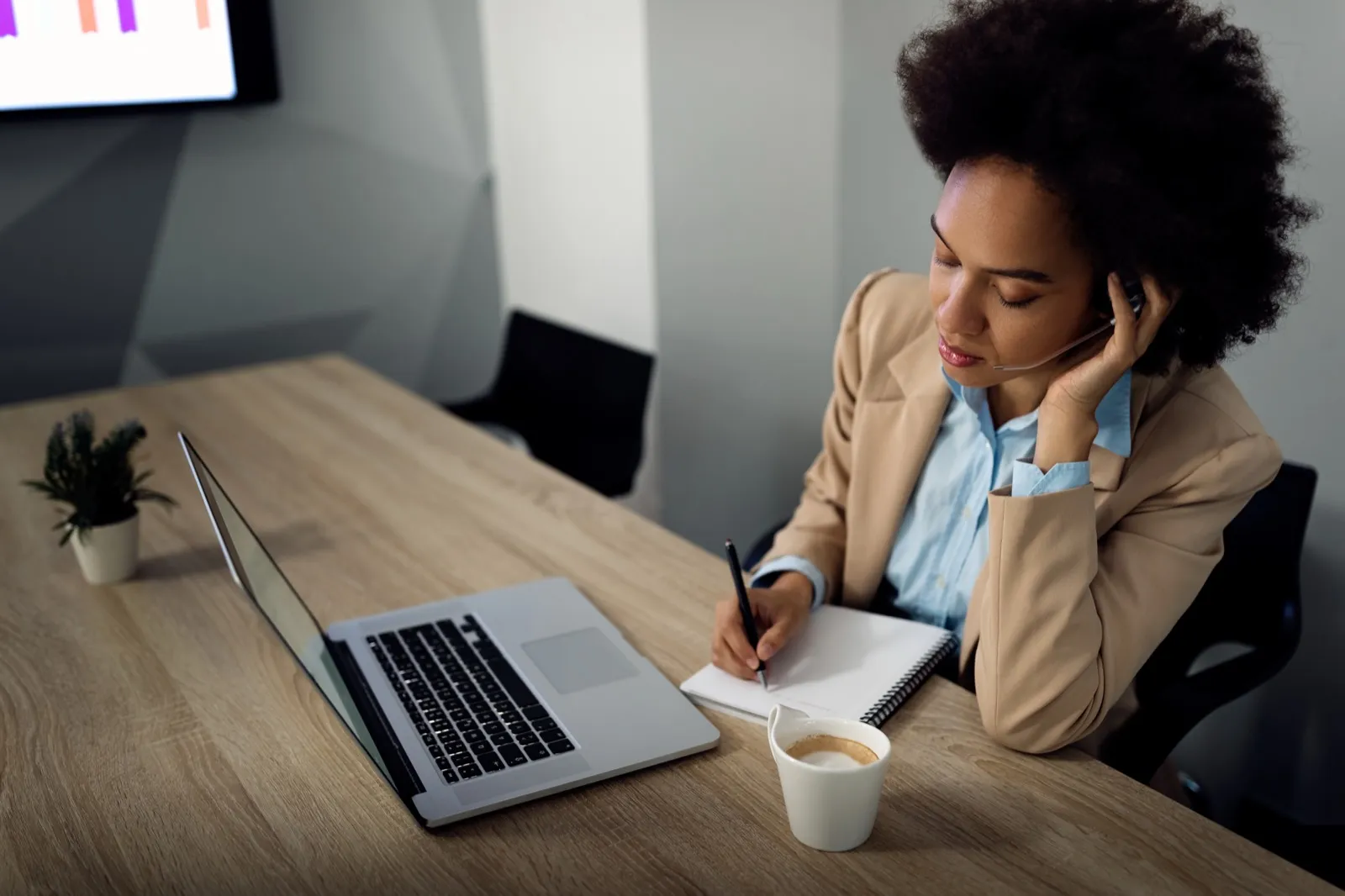 A woman writes notes in a pad beside an open laptop.