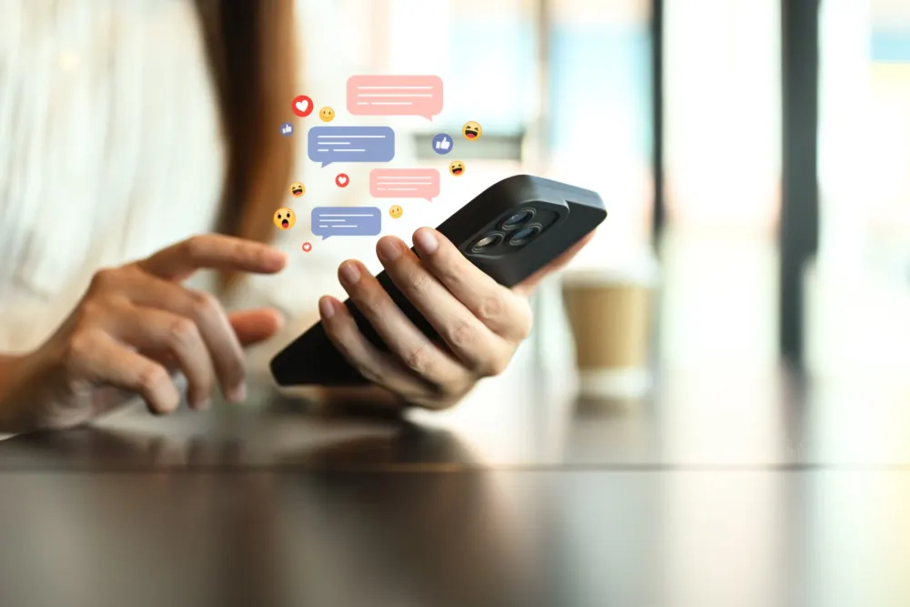 A hand holding a phone, with message bubbles and reactions rising out of the screen.