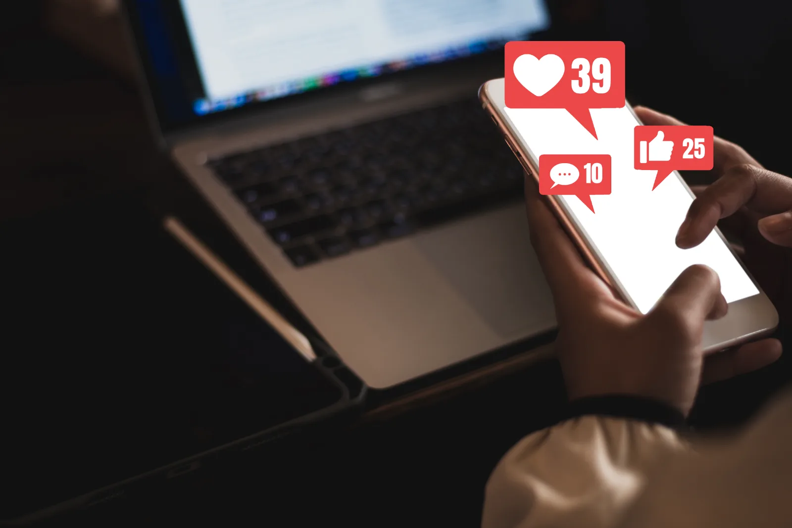 A hand holds a phone showing like, comment and message counts, above a laptop.