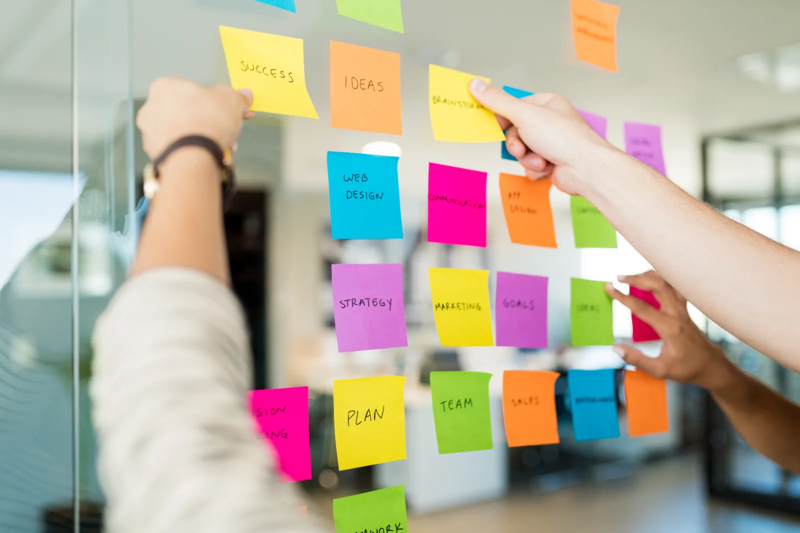 Hands arranging sticky notes labelled strategy, plan and goals on an office glass wall.