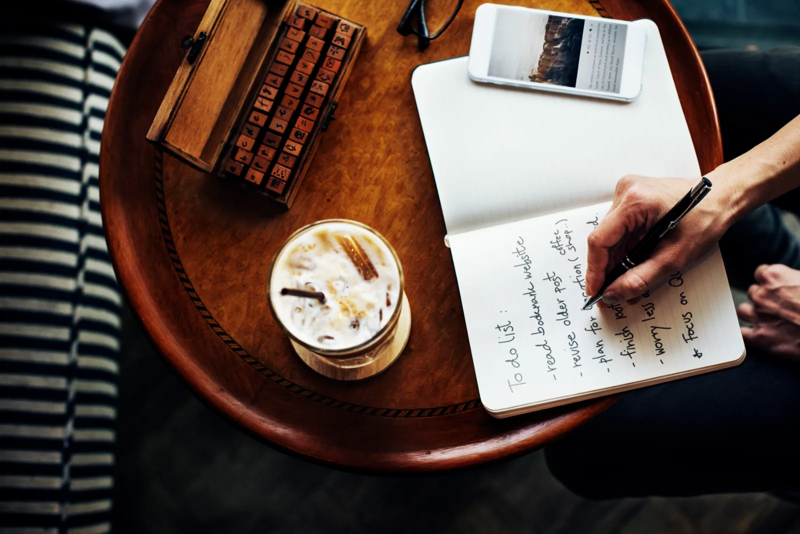 A hand writes a to-do list in a notebook beside an iced coffee.