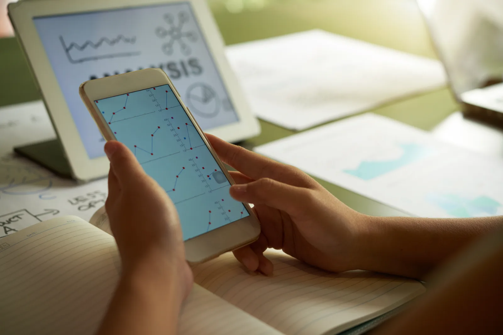 Hands compare charts on a phone against an analysis on a tablet, printed graphs beside them.